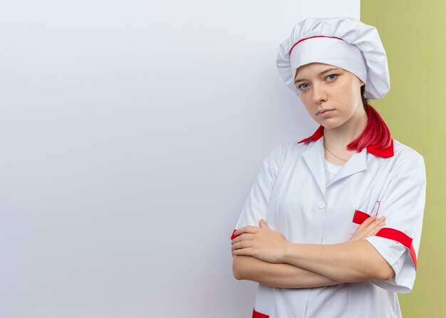 Jovem chef feminina loira confiante com uniforme de chef em frente a uma parede branca com os braços cruzados, isolados na parede verde