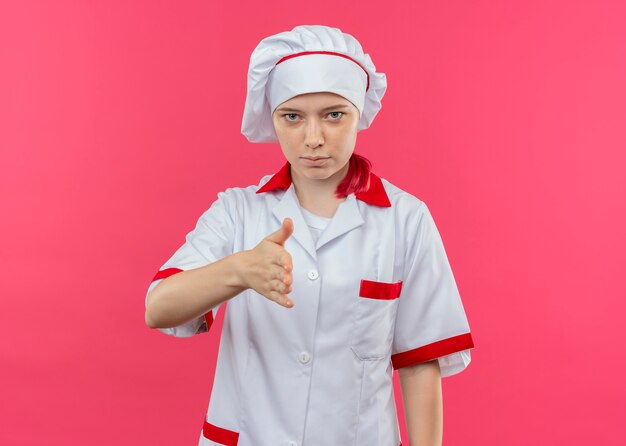 Jovem chef feminina loira confiante com uniforme de chef segurando a mão isolada na parede rosa