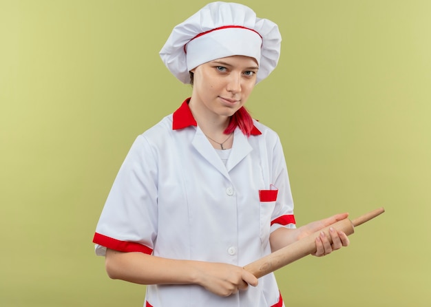 Jovem chef feminina loira confiante com uniforme de chef segurando o rolo de massa nas mãos e parece isolado na parede verde