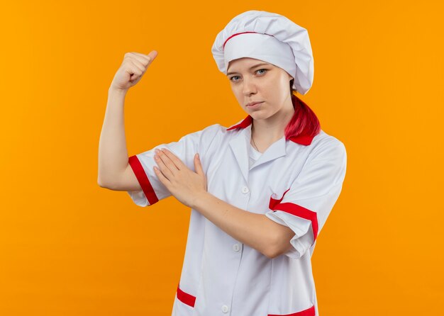 Jovem chef feminina loira confiante em tempo de uniforme de chef e coloca a mão no bíceps isolado na parede laranja