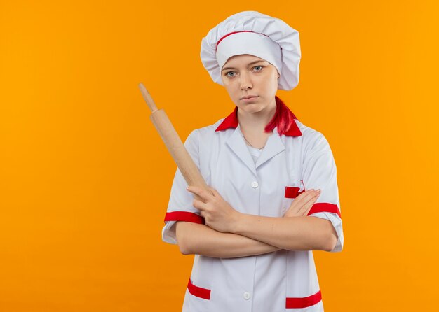 Jovem chef feminina loira confiante em uniforme de chef cruza os braços e segura o rolo de massa isolado na parede laranja