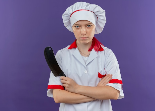 Jovem chef feminina loira confiante em uniforme de chef cruza os braços e segura uma faca isolada na parede violeta