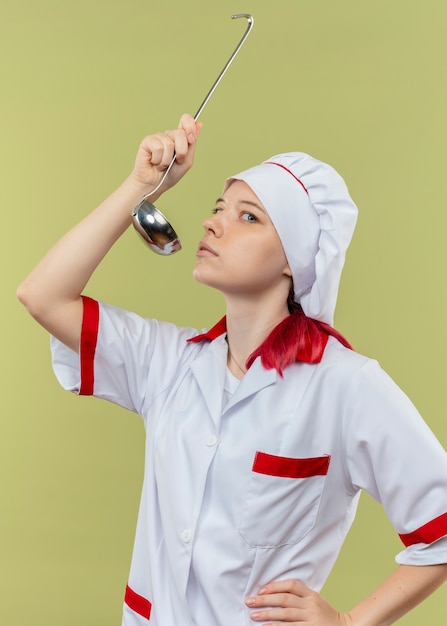Foto grátis jovem chef feminina loira confiante em uniforme de chef finge experimentar com uma concha isolada na parede verde