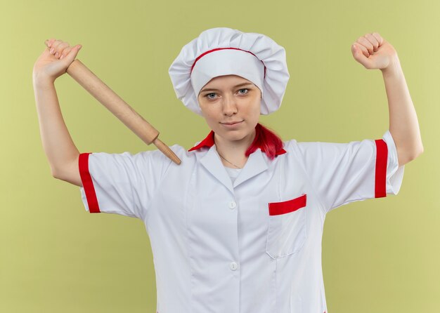 Jovem chef feminina loira confiante em uniforme de chef segura o rolo de massa e levanta o punho isolado na parede verde