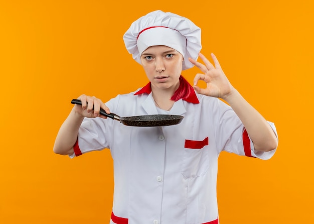 Jovem chef feminina loira confiante em uniforme de chef segurando uma frigideira e gesticulando com a mão bem isolada na parede laranja