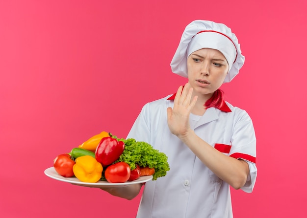 Jovem chef feminina loira irritada com uniforme de chef segura legumes no prato e finge empurrar com a mão isolada na parede rosa