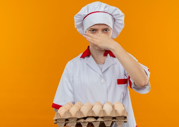 Jovem chef feminina loira irritada com uniforme de chef segura lote de ovos e fecha o nariz com a mão isolada na parede laranja