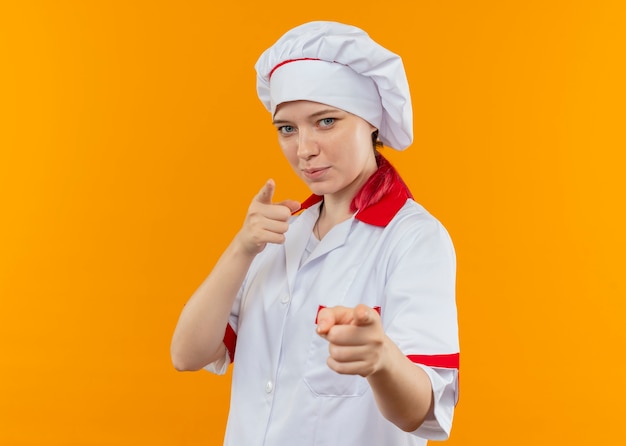 Jovem chef feminina loira satisfeita com o uniforme do chef aponta com as duas mãos isoladas na parede laranja