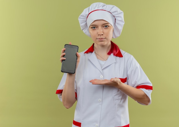 Jovem chef feminina loira satisfeita com uniforme de chef agarra e aponta com a mão no telefone isolado na parede verde