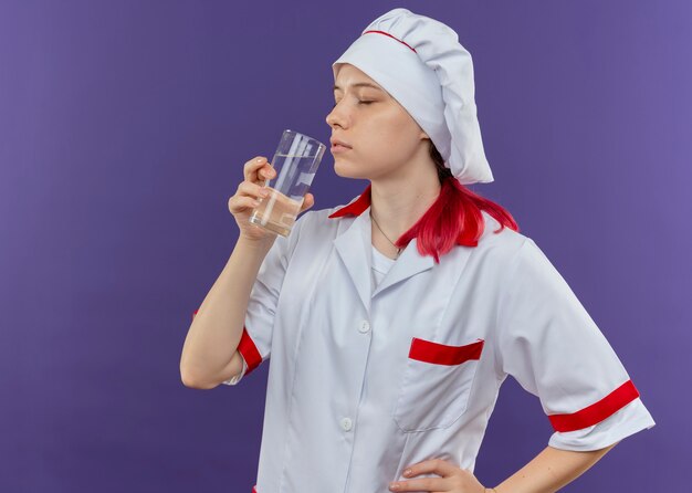 Jovem chef feminina loira satisfeita com uniforme de chef fingindo beber um copo de água isolado na parede violeta