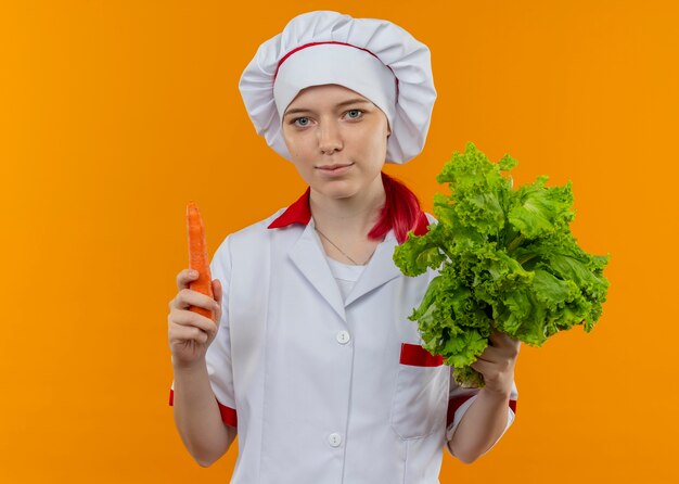 Jovem chef feminina loira satisfeita com uniforme de chef segurando salada e cenoura isoladas na parede laranja