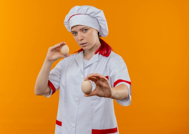 Jovem chef loira confiante com uniforme de chef segurando ovos isolados na parede laranja