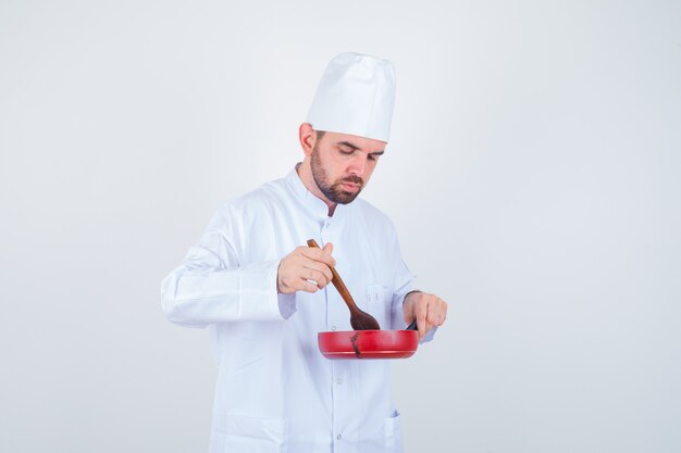 Jovem chef masculino em uniforme branco, mistura de refeição com colher de pau e olhando curioso, vista frontal.