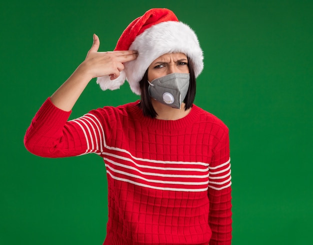 Foto grátis jovem cheia de chapéu de papai noel e máscara protetora fazendo gesto de suicídio isolado na parede verde