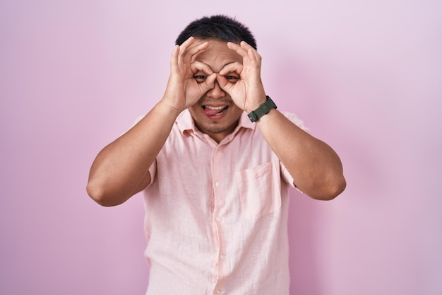 Foto grátis jovem chinês em pé sobre fundo rosa fazendo gesto ok como binóculos enfiando a língua para fora olhos olhando por entre os dedos expressão louca