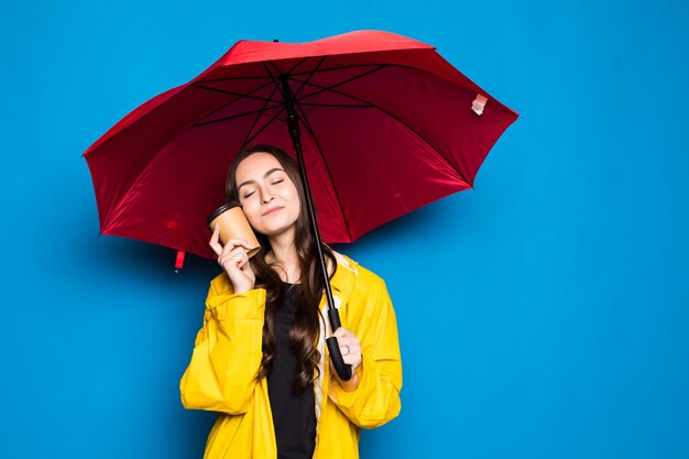 Jovem chinesa vestindo uma capa de chuva segurando um guarda-chuva sobre uma parede azul isolada muito feliz apontando com a mão e o dedo