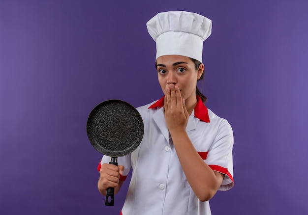 Foto grátis jovem chocada caucasiana cozinheira com uniforme de chef segura a frigideira e fecha a boca com a mão isolada na parede violeta com espaço de cópia