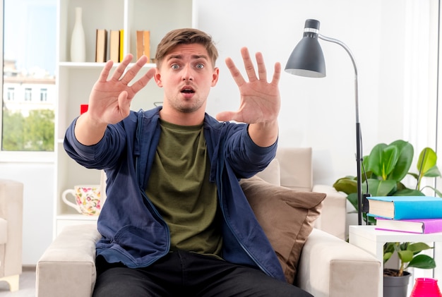 Foto grátis jovem chocado e bonito homem sentado na poltrona gesticulando nove com os dedos dentro da sala