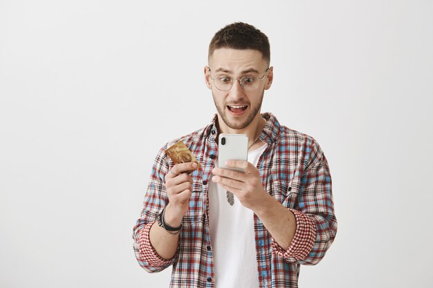 Jovem chocado e surpreso com óculos, posando com seu telefone e cartão