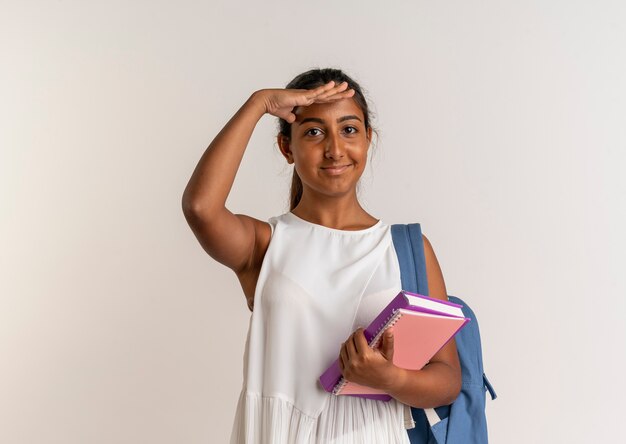 Jovem colegial satisfeita vestindo uma bolsa de costas segurando um caderno e olhando para a distância com a mão no branco
