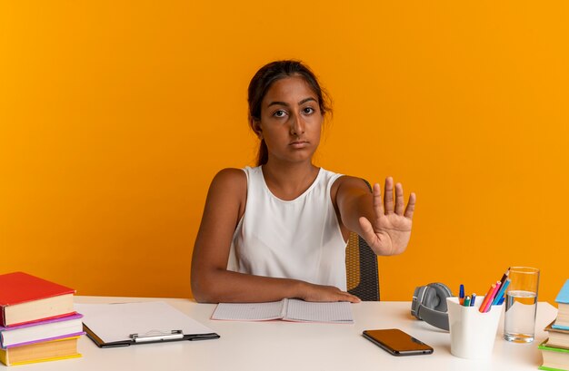 Jovem colegial sentada na mesa com as ferramentas da escola mostrando um gesto de pare