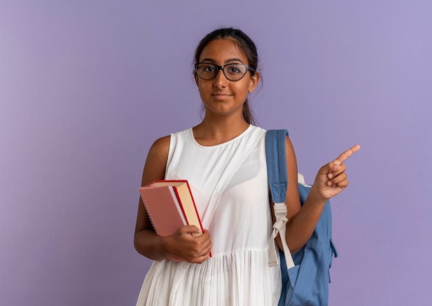 Foto grátis jovem colegial usando mochila e óculos segurando livros e apontando para o lado