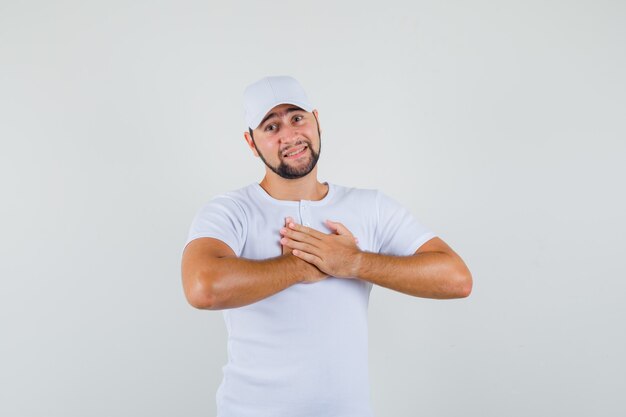 Jovem colocando as mãos no peito em t-shirt branca, boné e parecendo desconfortável, vista frontal.