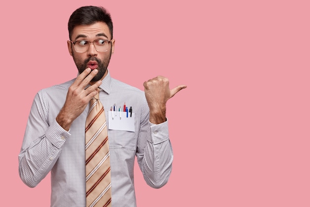 Jovem com a barba por fazer chocado cobre a boca, parece surpreso, vestido com roupas formais com gravata, aponta com o polegar para o espaço da cópia