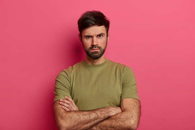 Jovem com a barba por fazer descontente franze a testa, olha com expressão severa e séria, mantém as mãos cruzadas sobre o corpo, ouve alguém, espera explicações, exige atenção, veste camiseta casual