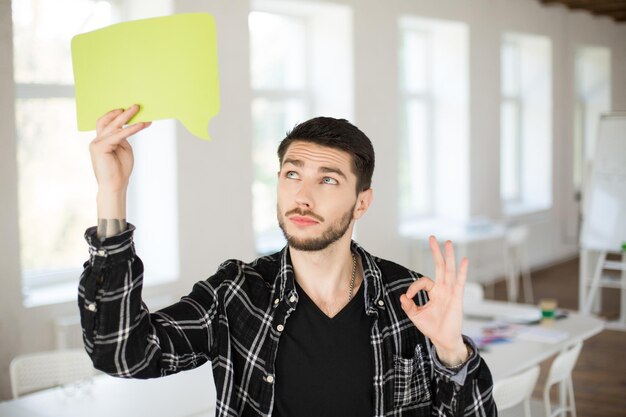 Jovem com barba olhando pensativamente no papel verde em forma de nuvem na mão perto da cabeça mostrando o gesto okey no escritório moderno