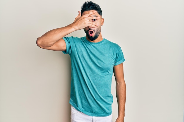 Jovem com barba vestindo roupas casuais espreitando em estado de choque cobrindo o rosto e os olhos com a mão olhando por entre os dedos com medo