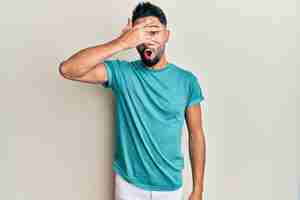 Foto grátis jovem com barba vestindo roupas casuais espreitando em estado de choque cobrindo o rosto e os olhos com a mão olhando por entre os dedos com medo