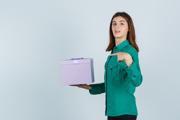 Jovem com blusa verde, calça preta segurando uma caixa de presente, apontando para ela e olhando alegre, vista frontal.