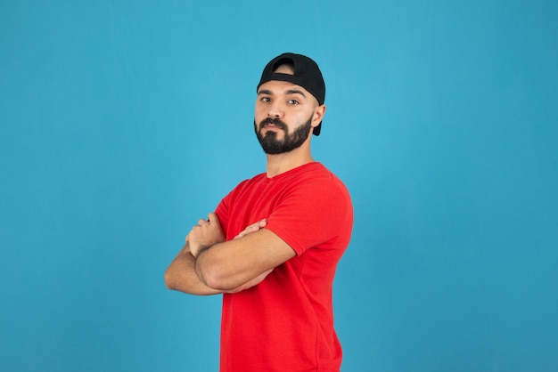 Jovem com boné vestindo camiseta vermelha em pé com os braços cruzados.