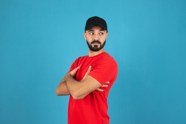 Jovem com boné vestindo camiseta vermelha em pé com os braços cruzados.