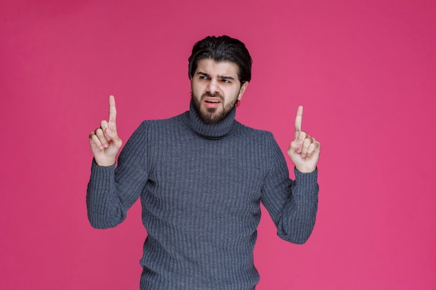 Jovem com cabelos longos e barba, mostrando ou apontando para algo com os dedos.