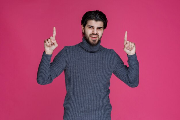 Jovem com cabelos longos e barba, mostrando ou apontando para algo com os dedos.