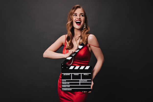Foto grátis jovem com cabelos ondulados e maquiagem linda posando com claquete mulher emocional feliz com vestido vermelho festivo sorrindo em fundo preto