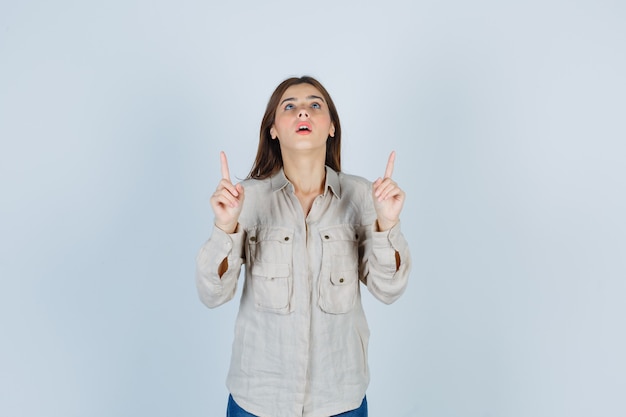 Jovem com camisa bege, jeans apontando para cima com o dedo indicador, olhando para cima e olhando a vista frontal, focada.