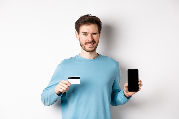 Jovem com camisa casual, mostrando a tela do smartphone vazia e cartão de crédito de plástico, piscando e sorrindo para a câmera, de pé no fundo branco.