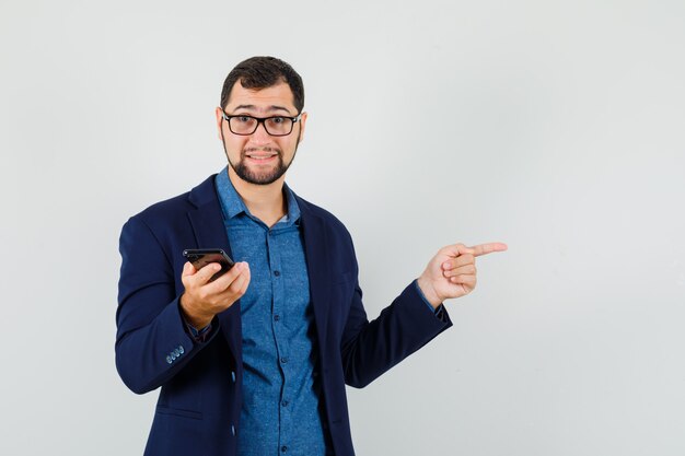 Jovem com camisa, jaqueta, segurando o telefone celular, apontando para a lateral, vista frontal.