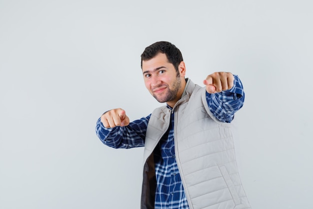 Jovem com camisa, jaqueta sem mangas, apontando para a frente e olhando alegre, vista frontal.