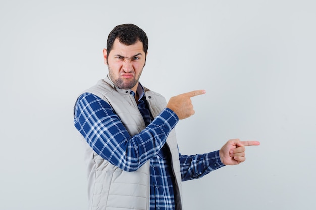 Jovem com camisa, jaqueta sem mangas apontando para o lado e parecendo descontente, vista frontal.