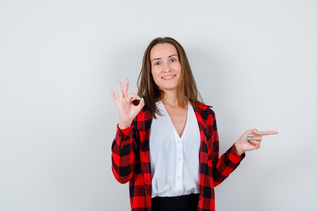 Jovem com camisa quadriculada, blusa mostrando o gesto de ok, apontando para o lado e parecendo feliz, vista frontal.