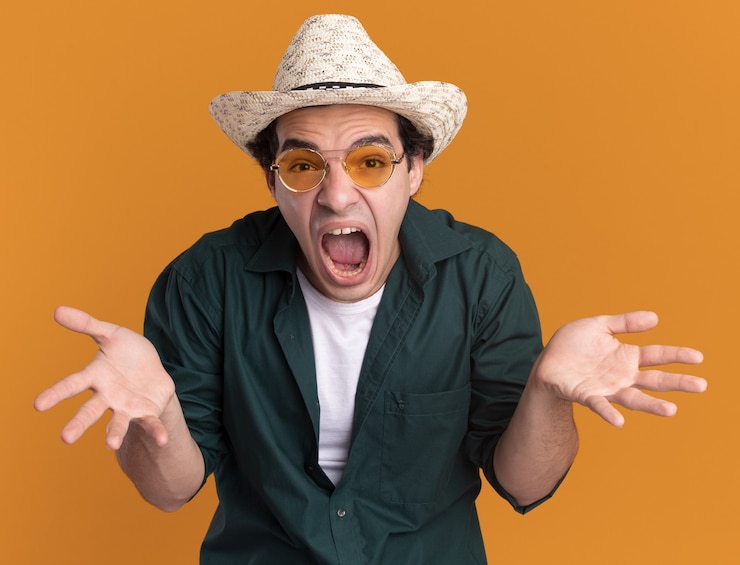 Foto grátis jovem com camisa verde e chapéu de verão, usando óculos, olhando para a frente, gritando e ficando louco, levantando as mãos em pé sobre a parede laranja