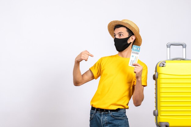 Jovem com camiseta amarela em pé perto da mala amarela segurando uma passagem e apontando para si mesmo