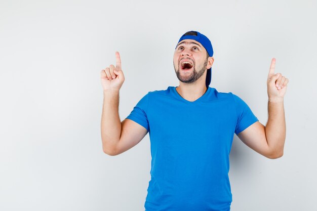 Jovem com camiseta azul e boné apontando para cima e parecendo feliz