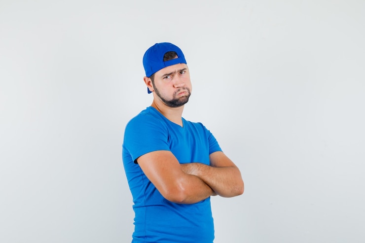 Foto grátis jovem com camiseta azul e boné em pé com os braços cruzados e parecendo desapontado