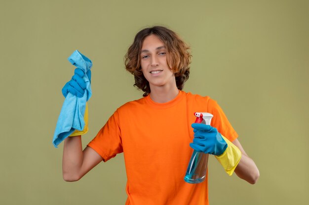 Jovem com camiseta laranja usando luvas de borracha segurando spray de limpeza e tapete olhando para a câmera sorrindo confiante em pé sobre fundo verde