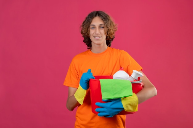 Jovem com camiseta laranja usando luvas de borracha, segurando um balde com ferramentas de limpeza, olhando para a câmera, sorrindo, positivo e feliz em pé sobre o fundo rosa